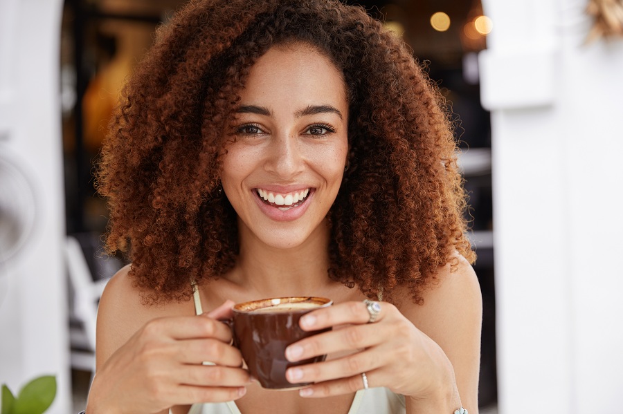 young African American female model has dark healthy skin, white teeth, drinks aromatic espresso, spends leisure time in coffee shop